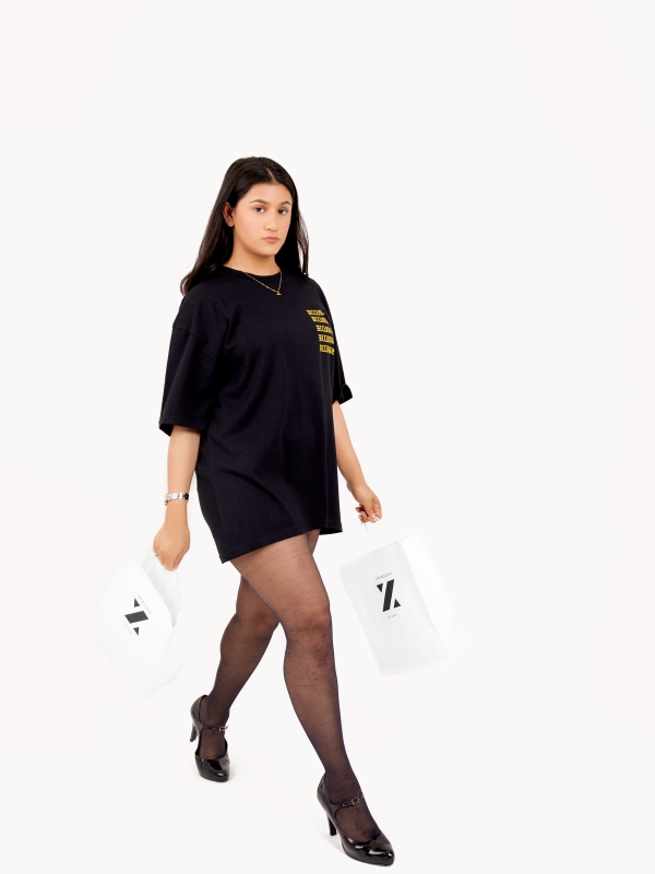 Woman In An Oversized Black T Shirt And Heels Walking While Carrying A White Shopping Bag.